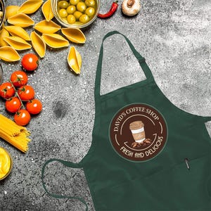 May include: Dark green apron with a circular logo for David's Coffee Shop, featuring a coffee cup and beans. The apron has a pocket and is surrounded by ingredients like tomatoes, pasta, and olives.