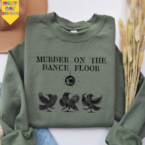 May include: An olive green sweatshirt with the text "MURDER ON THE DANCE FLOOR" above a graphic of three crows. A disco ball graphic is between the text and the crows. The sweatshirt is folded on a white surface.