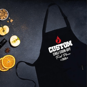 May include: Black apron with the text "CUSTOM CHILI COOK-OFF First Place *2026*" in white. A red flame graphic is above the text. The apron is on a dark surface with sliced oranges, apples, and a small bowl of brown sugar.