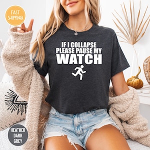 May include: Heather dark grey t-shirt with the text "IF I COLLAPSE PLEASE PAUSE MY WATCH" in white. A white graphic of a running person is below the text. The shirt is a crew neck style.