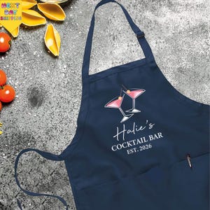 May include: Navy blue apron with a cocktail graphic and the text "Halie's COCKTAIL BAR EST. 2026". The apron has a pocket and adjustable neck strap. The background includes tomatoes and lemon wedges.
