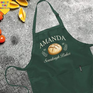 May include: A dark green apron with the name "AMANDA" in white script above a watercolor illustration of a loaf of bread. The text "Sourdough Baker" is below the bread illustration. A pen is in the pocket.