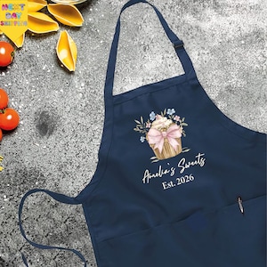 May include: Navy blue apron with a cupcake design and the text "Amelia's Sweets Est. 2026". The cupcake has a pink bow and floral accents. The apron has a pocket and adjustable neck strap.