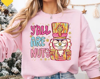 Op de afbeelding: Roze sweatshirt met de tekst "Y'all Are Nuts" en een kleurrijke notenkraker-afbeelding. De notenkraker draagt een hoge hoed en een goud-roze uniform. De sweatshirt heeft een ronde hals.