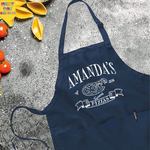 May include: Navy blue apron with the text "AMANDA'S Famous Pizzas" in white. The apron has a pocket and adjustable neck strap. The design includes a pizza graphic and the year 2026.