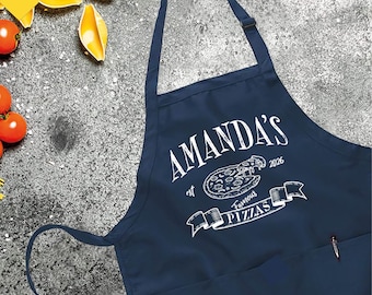 Puede incluir: Delantal azul marino con el texto blanco "AMANDA'S Famous Pizzas". El delantal tiene un bolsillo y una correa ajustable para el cuello. El diseño incluye un gráfico de pizza y el año 2026.