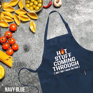 May include: Navy blue apron with the text "HOT STUFF COMING THROUGH" in white, with a flame graphic. The apron has a pocket and a pen in the pocket. The apron is surrounded by ingredients like tomatoes, pasta, and olives.