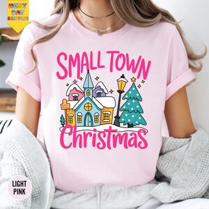 May include: Light pink t-shirt with the words "SMALL TOWN Christmas" in pink script. The graphic features a cartoon village scene with a church, houses, a lamppost, and a Christmas tree. The shirt has short sleeves.