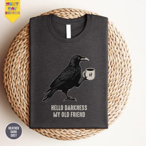 May include: Dark heather gray t-shirt featuring a black raven holding a coffee cup. The raven is illustrated with detailed feathers and a curved beak. The text "HELLO DARKNESS MY OLD FRIEND" is printed below the raven.