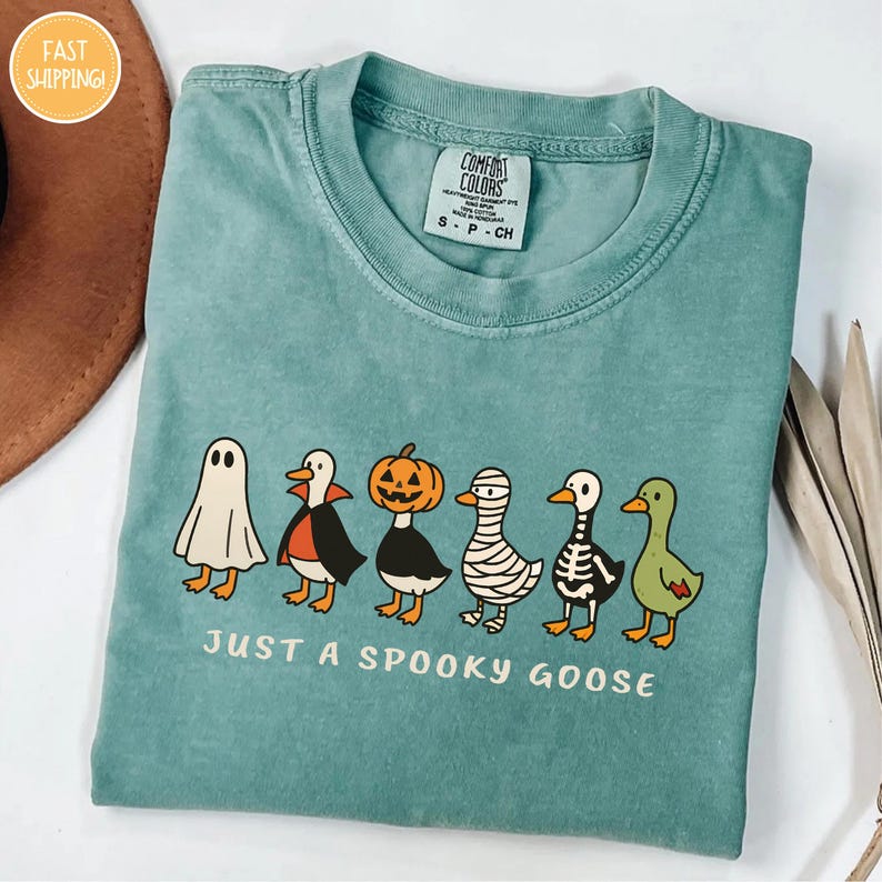 A turquoise t-shirt with a Halloween-themed graphic design featuring a skeleton goose, a jack-o-lantern, and a ghost. The text below the graphic says "Just a spooky goose".