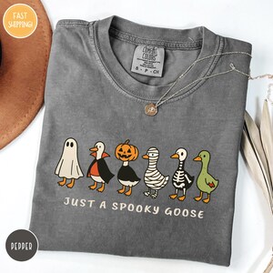 A gray t-shirt with a Halloween-themed graphic design, featuring a skeleton goose, a jack-o-lantern, a ghost, and a pepper. The text on the t-shirt says "Just a Spooky Goose".