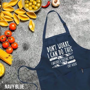 May include: Navy blue apron with the text "DON'T WORRY, I CAN DO THIS. I WATCHED A YOUTUBE VIDEO - CHEF ALBERT" in white. The apron has a pocket and a pen. Pasta, tomatoes, and olives are in the background.