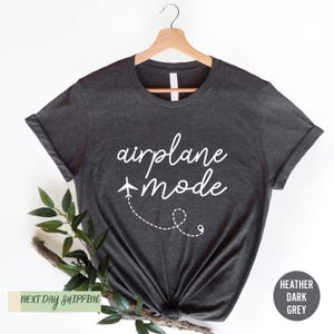 May include: Heather dark grey t-shirt featuring the text 'airplane mode' in a stylish script font. A small airplane icon with a dotted line trail is included in the design. The shirt is displayed on a hanger, showcasing its relaxed fit and soft fabric.  Next day shipping is offered.  The overall theme is travel and relaxation.