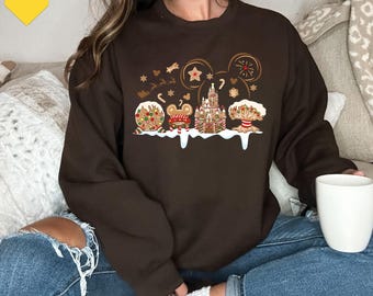 Op de afbeelding: Een donkerbruin sweatshirt met lange mouwen en een grafische print van vakantie-iconen in peperkoekstijl, waaronder een kasteel, Mickey Mouse-oren en feestelijke lekkernijen op een besneeuwde witte basis met verspreide gouden sterren.