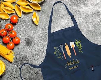 May include: Navy blue apron with a pocket and adjustable neck strap. The apron features a design with a whisk, rolling pin, and spatula, along with the text "Adela's BAKERY" in gold lettering. The apron is surrounded by ingredients like tomatoes and pasta.