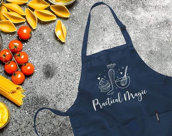 May include: Navy blue apron with a white graphic of a cupcake, whisk, and cactus, with the words "Practical Magic." The apron has a pocket and adjustable neck strap. Various food items surround the apron, including tomatoes, pasta, and olives.