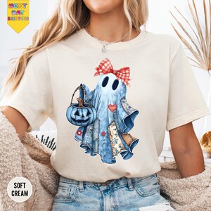 May include: Cream-colored t-shirt featuring a whimsical ghost design. The ghost, in shades of blue, is adorned with a red gingham bow and holds a blue pumpkin basket. Patches of floral and patchwork fabric add detail. The shirt has the words "Soft Cream" printed on it.