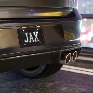 Op de afbeelding: Close-up van de achterkant van een grijze auto met een zwart kenteken met de tekst "JAX" in witte letters met een turquoise omlijning. Verchroomde uitlaten zijn ook zichtbaar.