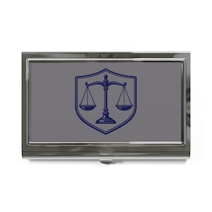 May include: A silver-toned business card holder with a gray center panel. The panel features a blue shield with a scale emblem, commonly associated with law and justice. The card holder has a shiny, reflective finish.