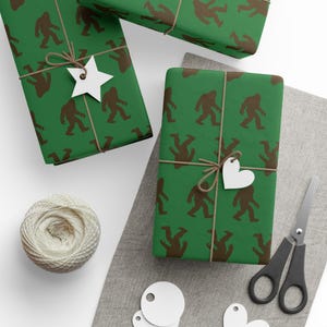 May include: Green wrapping paper featuring a repeating brown Bigfoot silhouette pattern. The packages are tied with brown twine and adorned with white star and heart-shaped gift tags. A ball of white twine and black scissors are also present.
