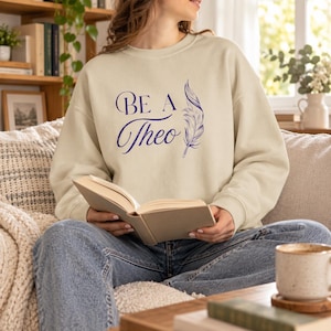 May include: A beige long-sleeve crewneck sweatshirt featuring dark blue text that reads "BE A Theo" alongside a detailed illustration of a single feather. The garment has a ribbed collar, cuffs, and hem, providing a classic fit and appearance.