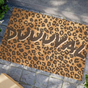 May include: A rectangular doormat with a leopard print pattern in shades of brown and tan. The word "DUVAL" is printed across the mat in a bold, stylized font. The mat is placed on a stone surface.