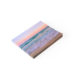 May include: A canvas print of a beach scene with a pink and orange sunset over the ocean. The waves are crashing on the sandy shore.