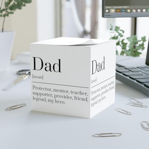 以下が含まれることがあります： 白い立方体型のメモ帳で、両面に黒字で「Dad」と印刷されています。「Protector, mentor, teacher, supporter, provider, friend, legend, my hero」と定義されています。メモ帳の周りには、いくつかの銀色のクリップが散らばっています。
