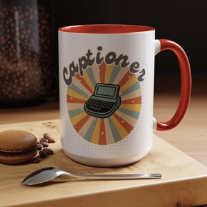 May include: White ceramic mug with a red interior and handle. The mug features a retro design with the word "captioner" and a vintage computer graphic on a colorful sunburst background. A spoon and a pastry are on a wooden surface.