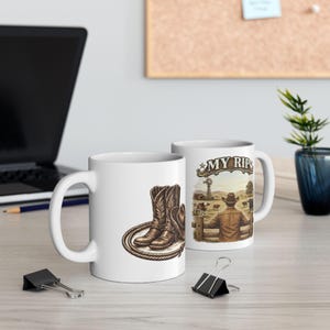 May include: Two white ceramic mugs featuring western-themed illustrations. One mug displays a pair of brown cowboy boots and a coiled lasso. The other mug shows a person in a cowboy hat looking over a wooden fence at a farm with the text MY RIP.