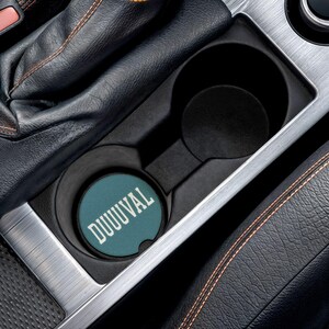 May include: A teal car coaster with the word "DUUUVAL" in white lettering sits in a car's cup holder. The cup holder is black and silver, and the surrounding interior is black leather with orange stitching.