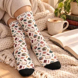 Könnte beinhalten: Ein Paar weiße Socken mit einem sich wiederholenden Muster aus bunten Büchern und Federn. Die Socken haben einfarbige schwarze Zehenbereiche und sind mit Illustrationen von Bücherstapeln, aufgeschlagenen Büchern sowie Federn in Rot, Grün und Blau verziert.