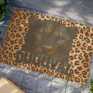 May include: A rectangular doormat with a leopard print background and a large image of a panther's face. The word "DUUUVAL" is printed in green letters across the bottom of the image.