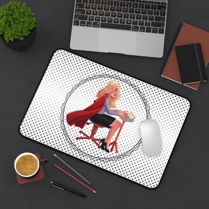 Puede incluir: Una alfombrilla de escritorio blanca con un patrón de lunares blanco y negro. Un marco circular blanco y negro rodea una ilustración de dibujos animados de una mujer con cabello rubio que lleva una capa roja y está sentada en una silla mientras escribe en una computadora portátil. Un ratón de computadora blanco está en la alfombrilla.