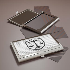 May include: A silver business card holder with a black and white scales of justice emblem. The holder is rectangular with a hinged lid and a dotted pattern on the front. Several business cards are visible.