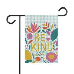Könnte beinhalten: Eine blaue und weiße Gingham-Gartenflagge mit einem floralen Design und dem Text "Be Kind" in goldenen Buchstaben.