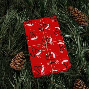 May include: A wrapped gift with red paper featuring a pattern of white Santa hats, black stethoscopes, and snowflakes. The gift is tied with twine and rests on a bed of green pine needles and pine cones.