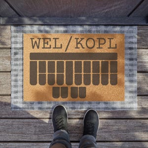 Puede incluir: Una alfombra rectangular con el texto "WEL/KOPL" en letras marrones. Debajo del texto hay un diseño de teclado marrón. La alfombra tiene un centro beige y un borde a cuadros gris y blanco. Se ven zapatos negros.