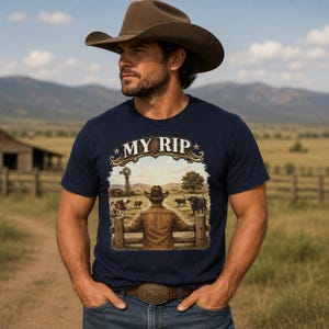 May include: A navy blue t-shirt featuring a graphic print of a person in a cowboy hat leaning on a wooden fence, looking out at a field with cattle and a windmill. The text "MY RIP" is printed in a decorative font above the scene.