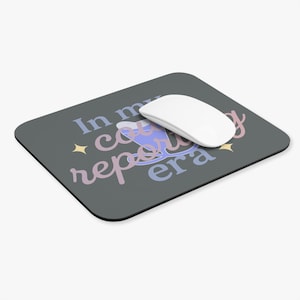 Puede incluir: Alfombrilla de ratón rectangular gris oscuro con el texto "In my country reporting era" en azul, rosa y morado. Un ratón de ordenador blanco descansa sobre la almohadilla.