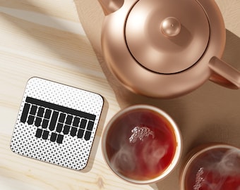 May include: A white coaster with a black keyboard design. The coaster is on a wooden surface with a copper teapot and two cups of tea.