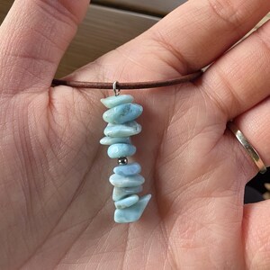 May include: A necklace featuring a pendant made of light blue, irregularly shaped stones. The stones are strung together with a silver bead and suspended from a brown leather cord. The necklace is held in a hand.