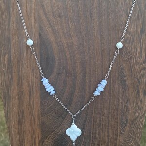 May include: A silver chain necklace featuring a four-leaf clover pendant and a teardrop charm. The necklace includes light blue and white beads. The necklace is displayed on a wooden surface.