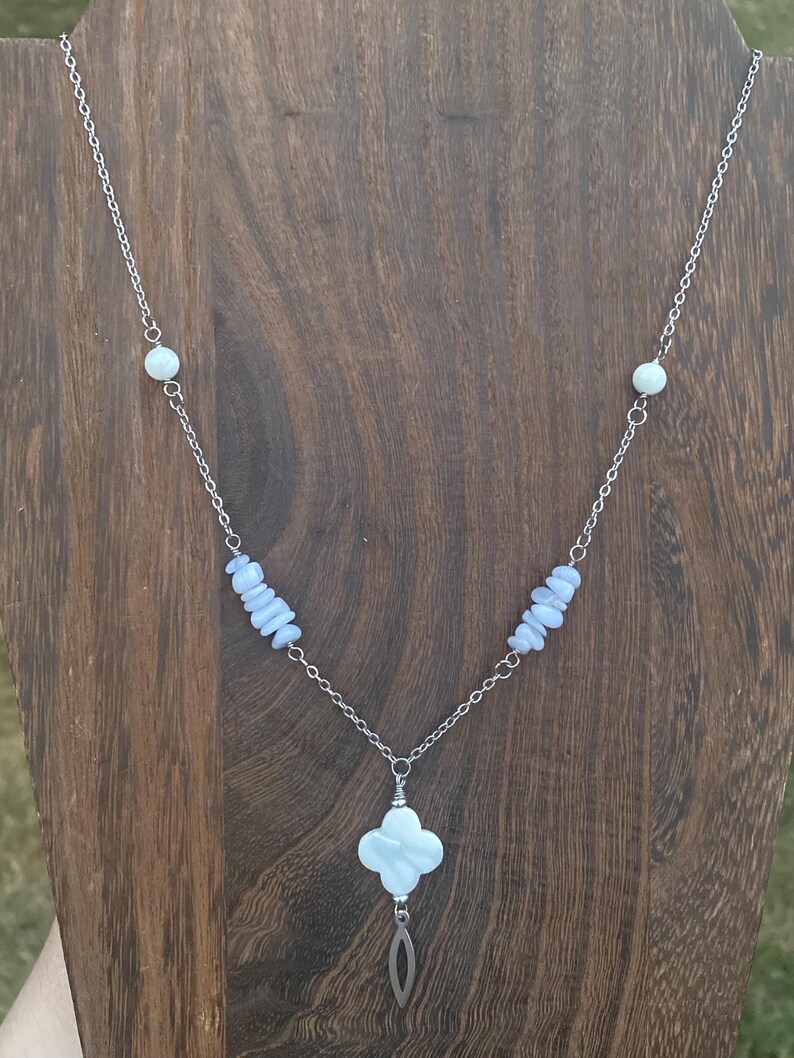 May include: A silver chain necklace featuring a four-leaf clover pendant and a teardrop charm. The necklace includes light blue and white beads. The necklace is displayed on a wooden surface.