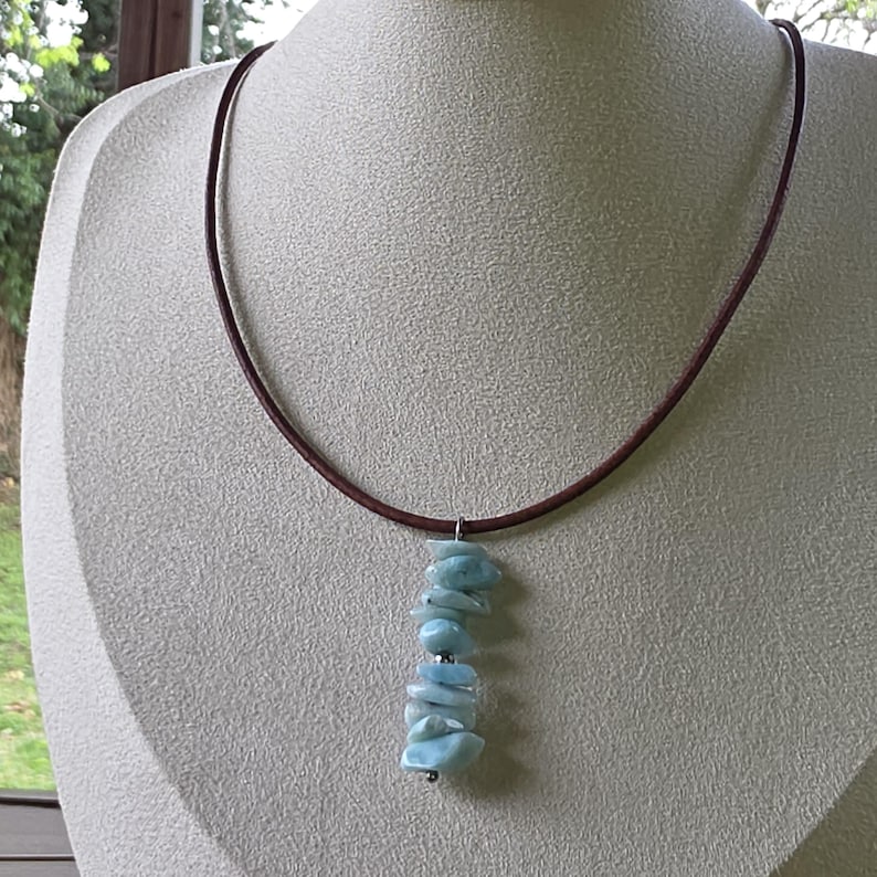May include: A brown leather necklace with a pendant of light blue, irregular-shaped stones. The stones are strung together with a silver-colored wire. The necklace is displayed on a beige mannequin.