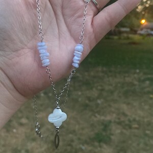 May include: A silver-toned necklace featuring a four-leaf clover pendant and a chain with blue and white stone accents. The necklace is displayed against a blurred outdoor background, showcasing the jewelry's delicate design.