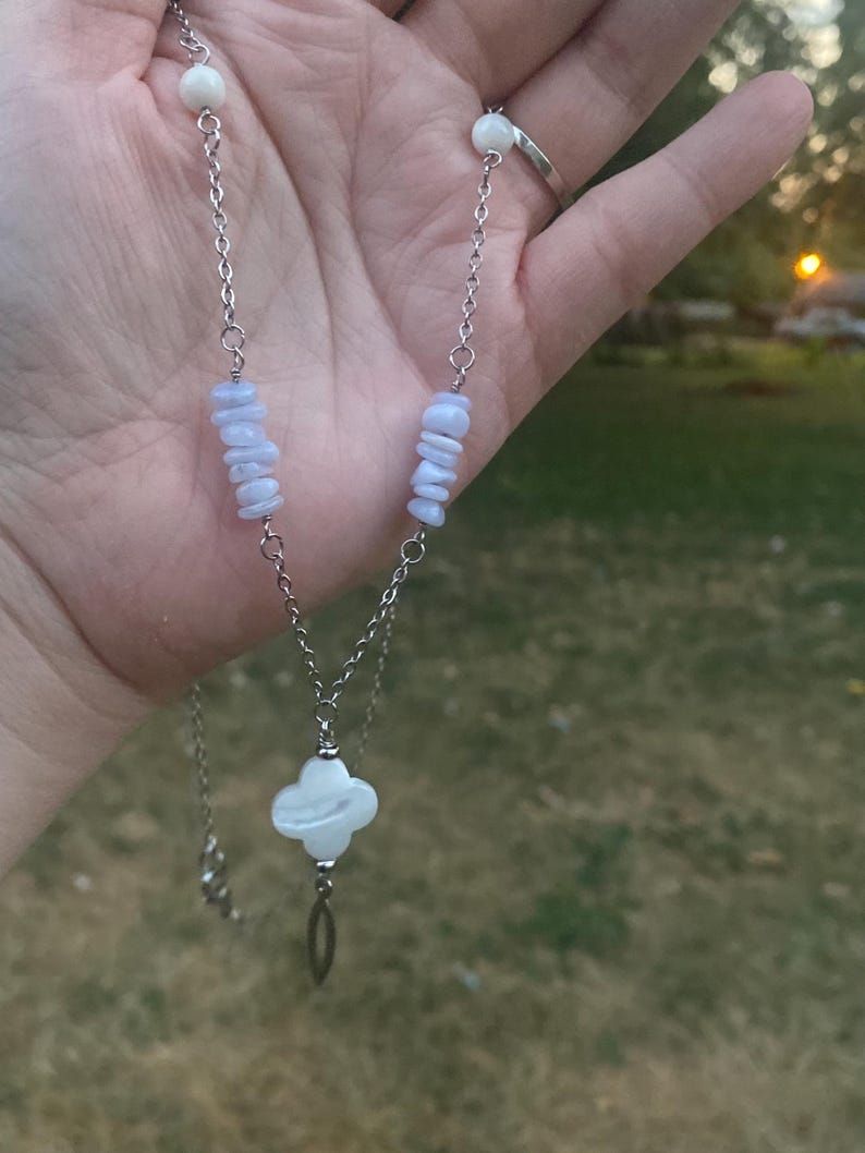 May include: A silver-toned necklace featuring a four-leaf clover pendant and a chain with blue and white stone accents. The necklace is displayed against a blurred outdoor background, showcasing the jewelry's delicate design.
