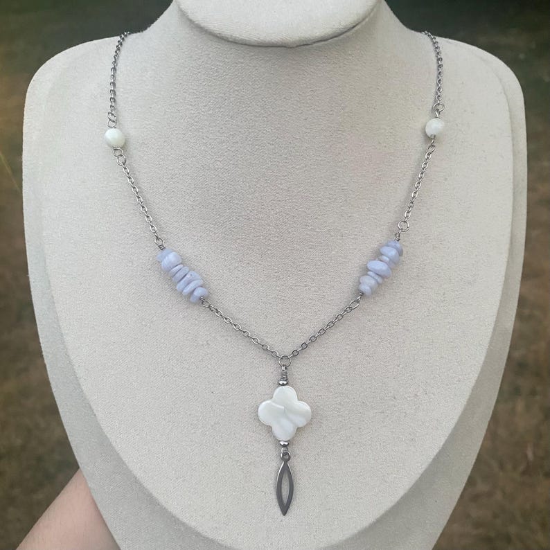 May include: A silver-toned necklace featuring a four-leaf clover pendant with a teardrop charm. The necklace has light blue and white beads, and a delicate chain. The jewelry is displayed on a mannequin.