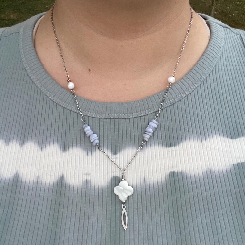 May include: Silver-toned necklace featuring a four-leaf clover pendant with a dangling teardrop shape. The necklace includes blue-gray stone beads and white pearl accents. The chain is delicate and hangs on a person wearing a light blue and white tie-dye shirt.