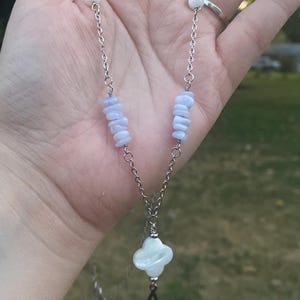 May include: A silver chain necklace featuring blue-grey stone chips and white beads. The pendant is a four-leaf clover shape with a dangling black teardrop. The necklace is held in a hand.